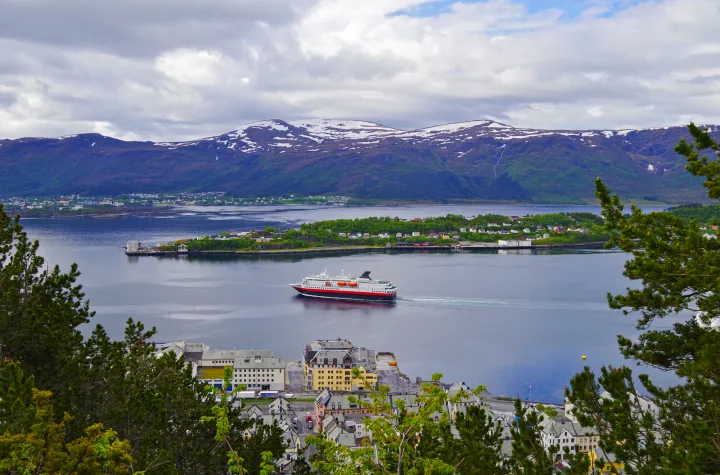 Schiff fährt auf einem Fjord, umgeben von Bergen und einem Küstendorf.